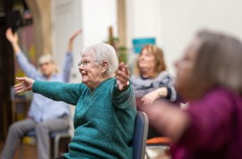 An image of a group of older people doing exercise on chairs in a room.